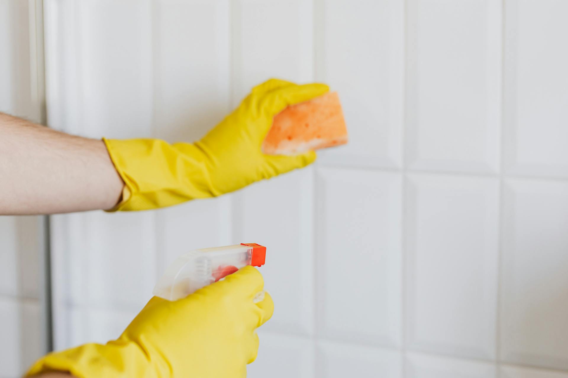 A person wearing yellow gloves cleans bathroom tiles with a sponge and spray bottle.