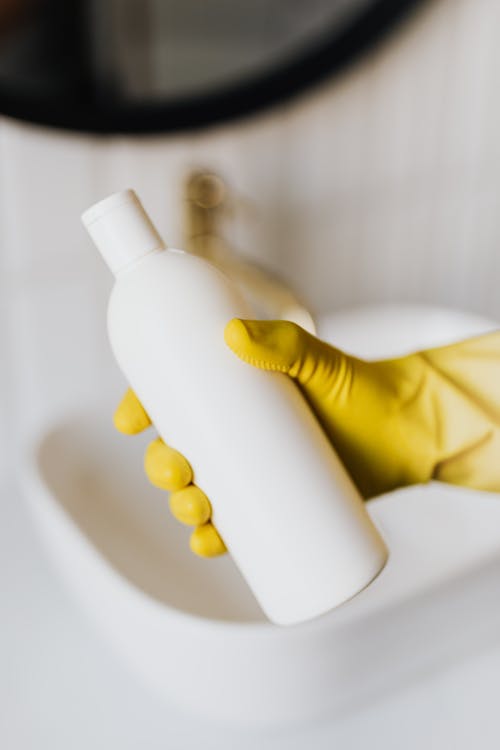 Crop anonymous person in yellow protective gloves demonstrating blank bottle of cosmetic product against sink in bathroom