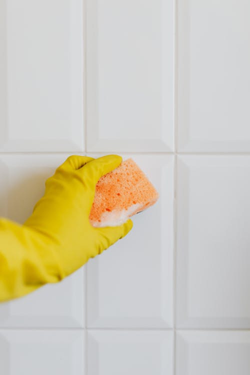 Crop anonymous person in yellow cleaning glove wiping white tile wall with wet orange cleaning sponge
