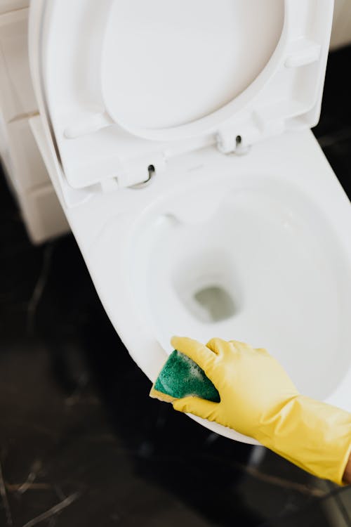 Free From above crop unrecognizable cleaner in rubber yellow gloves using sponge to wipe off toilet bowl in restroom Stock Photo