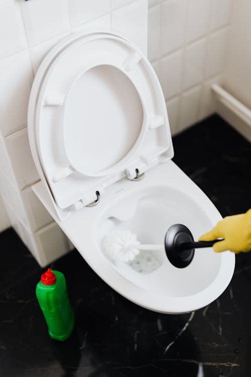 From above crop faceless person in yellow rubber cleaning gloves removing stains in toilet bowl with brush and detergent in washroom