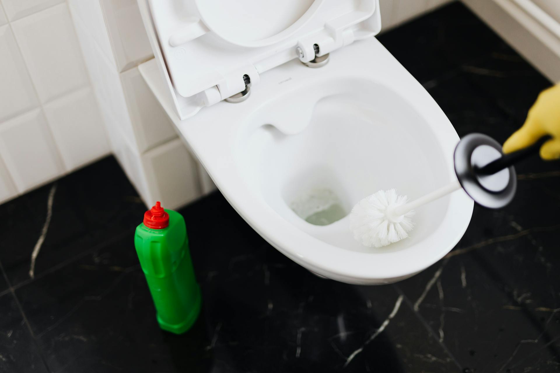 From above of crop anonymous person in rubber gloves using liquid toilet sanitizer and brush to clean toilet bowl in bathroom