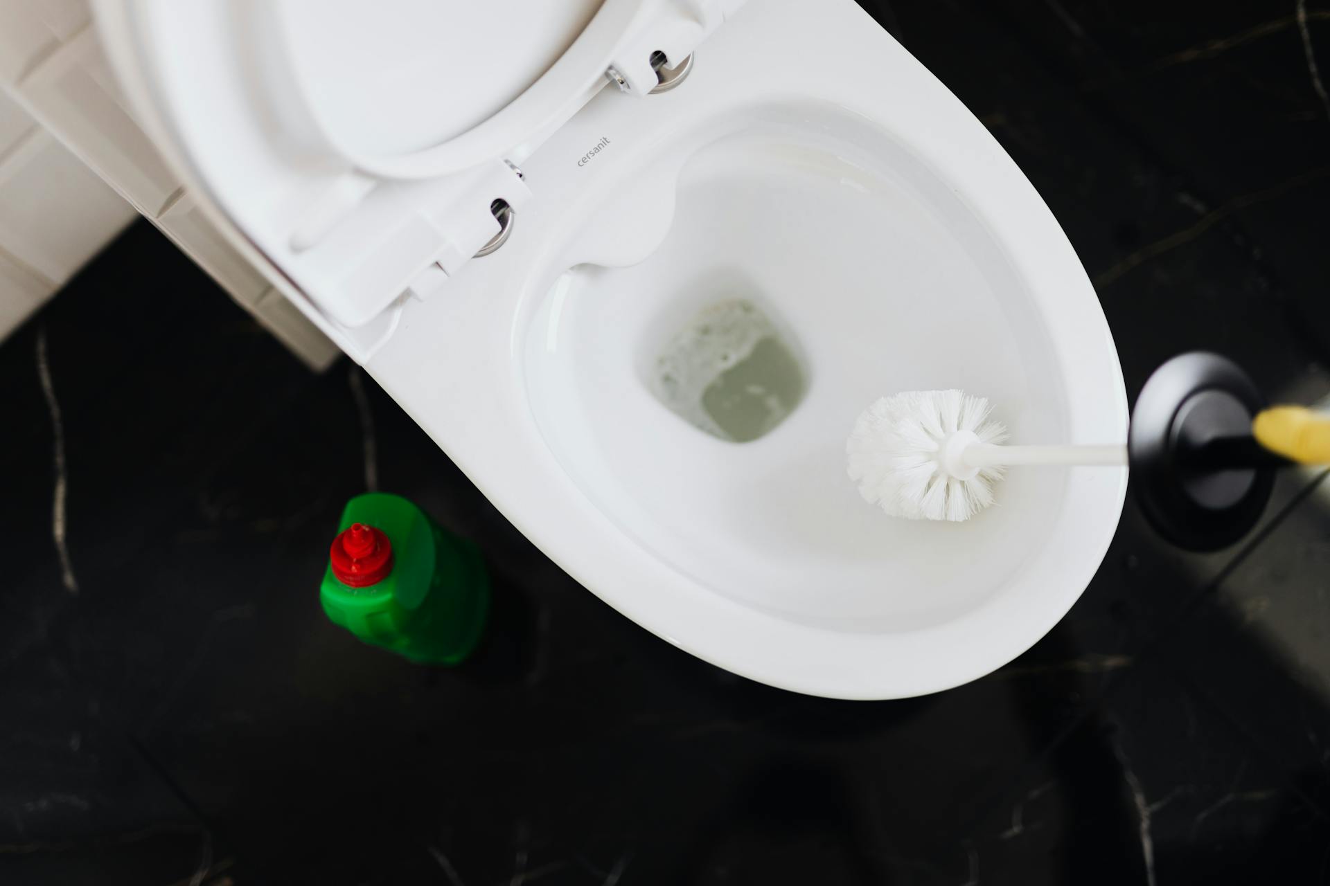 From above crop anonymous person in yellow gloves cleaning toilet bowl with toilet brush and liquid detergent in green plastic bottle