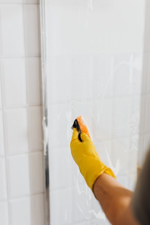 Crop faceless person in yellow rubber protective glove using detergent and sponge for cleaning walk in shower glass