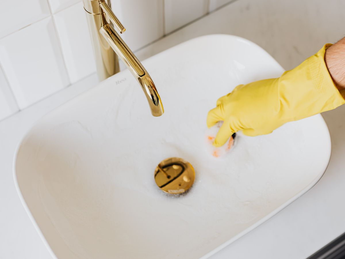 A person in yellow rubber gloves scrubbing a sink with a sponge and detergent