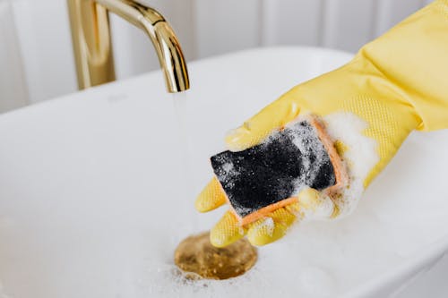 Free Crop faceless person in yellow latex protective glove using detergent and sponge while washing surface of white sink in bathroom Stock Photo