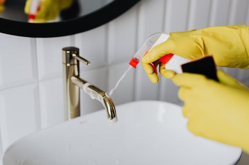 From above anonymous housekeeper in yellow rubber gloves spraying cleaner to chrome water faucet in bathroom