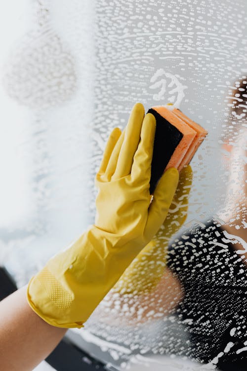 Faceless housekeeper in yellow rubber gloves cleaning mirror from foam by sponge in bathroom