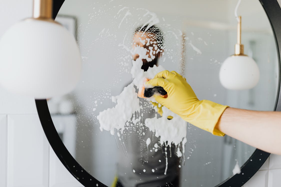Anonymous male housekeeper in yellow rubber gloves wiping foam from round mirror in bathroom