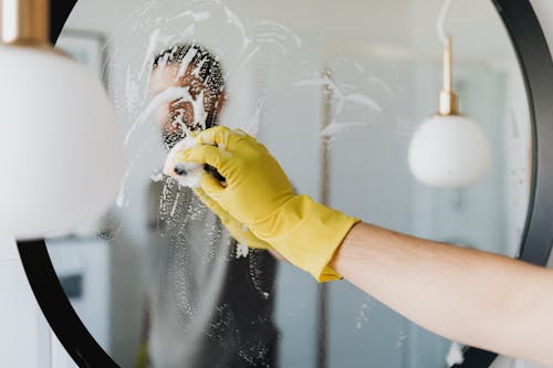 Anonymous male in yellow rubber gloves wiping foam from round mirror in bathroom