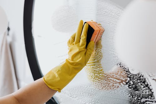 Free Anonymous person in yellow rubber gloves using simple sponge for cleaning mirror in bathroom Stock Photo