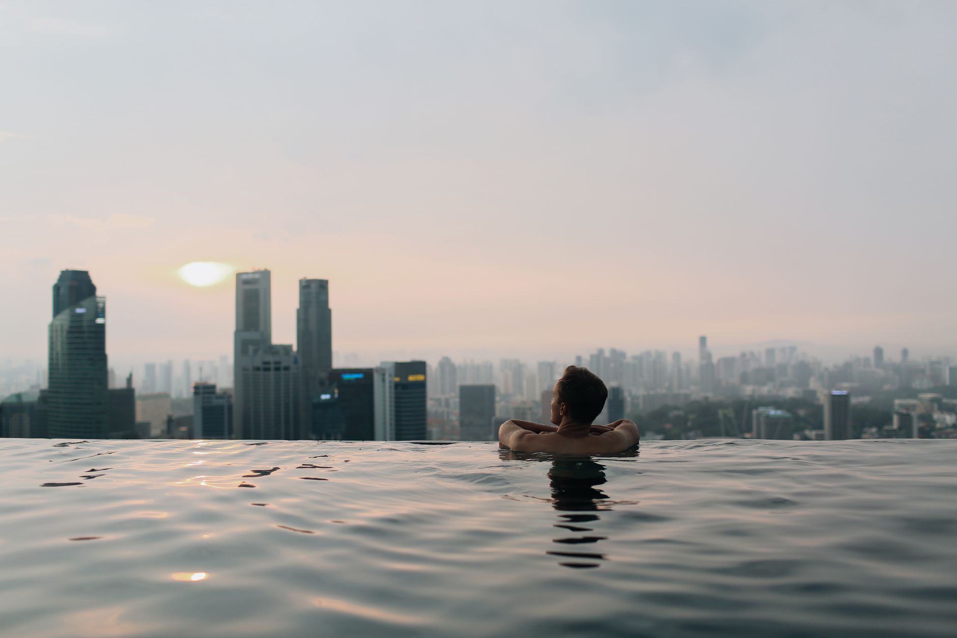 A man relaxes in an infinity pool with a breathtaking view of the Singapore city skyline at sunset.