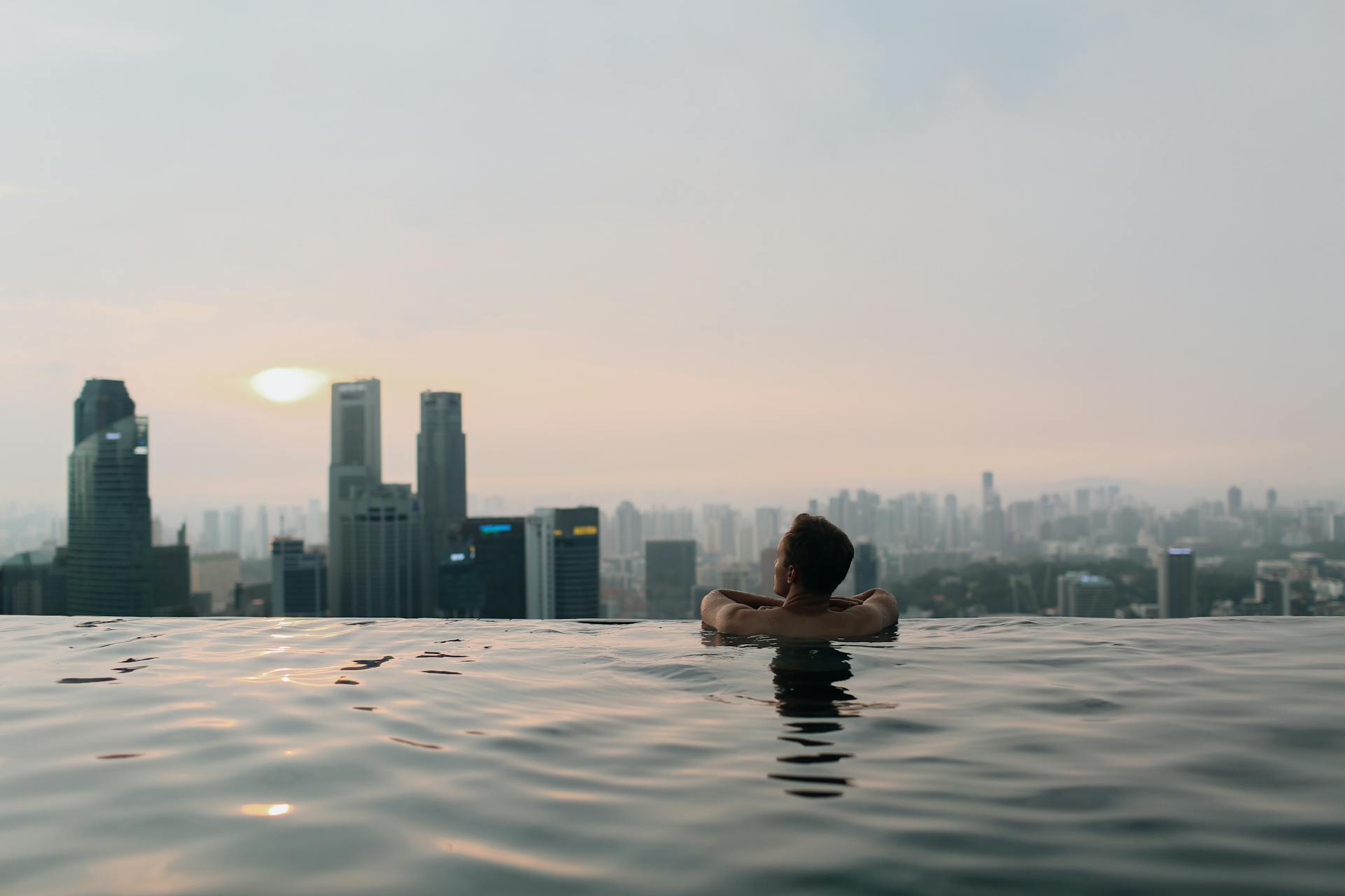 A man relaxes in an infinity pool with a breathtaking view of the Singapore city skyline at sunset.