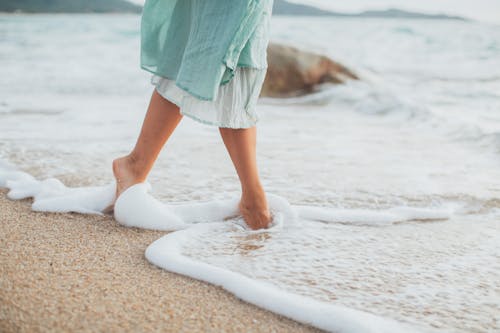 Free Unrecognizable crop barefooted female tourist in light dress walking in foamy waves of picturesque sea on sandy beach Stock Photo