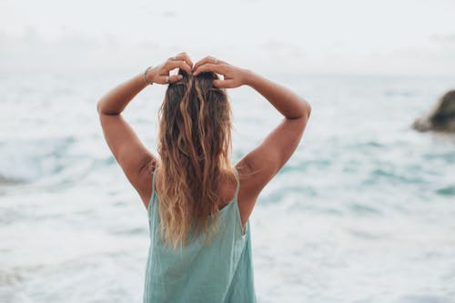 Back view of unrecognizable female in light top admiring powerful waving ocean with raised arms during summer holidays