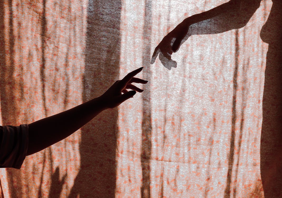 Free Crop unrecognizable man and woman hands reaching out to each other through translucent curtain on sunny day Stock Photo
