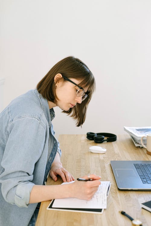 Free Focused young woman taking notes in a cozy home office setting with laptop and stationery. Stock Photo