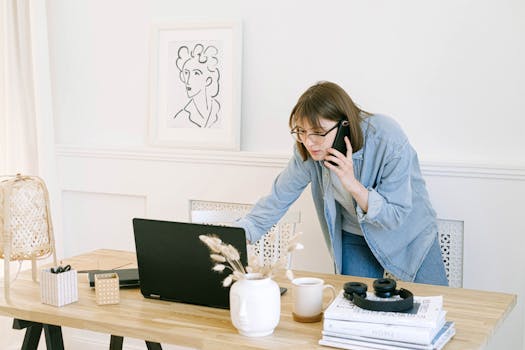 Woman multitasking with laptop and phone in modern home office setting.