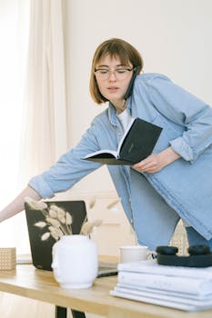 A young woman multitasks with a phone call and notebook in a casual home office setting.
