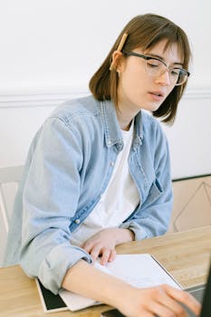 Young woman in casual attire writing notes at a home office desk, concentrating intently.