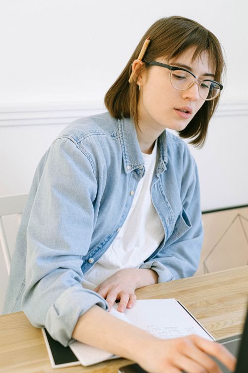 Free Young woman in casual attire writing notes at a home office desk, concentrating intently. Stock Photo