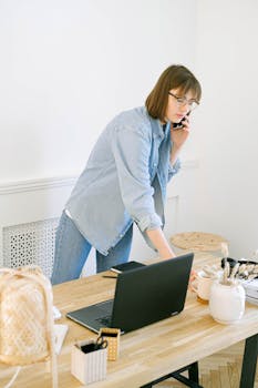 Woman multitasking with phone and laptop in bright home office setting.
