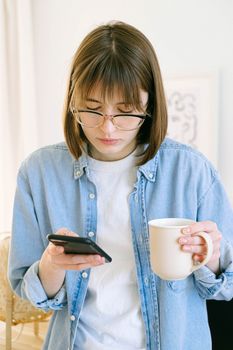 一名戴眼镜的年轻女子在室内拿着马克杯看手机