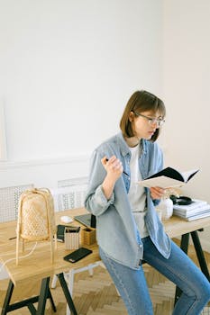 Young woman in glasses reading and taking notes in a casual home office setting with wooden decor.