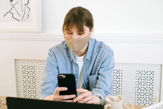 Woman wearing a face mask, working remotely from home using a smartphone and laptop.
