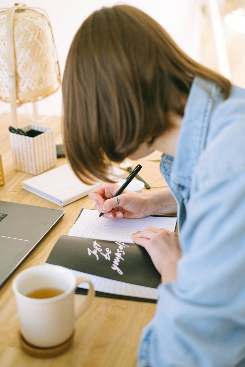 Woman writing notes at a desk with coffee and laptop