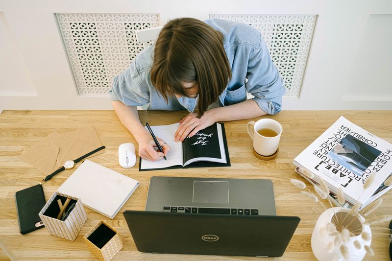 Overhead view of a desk where someone writes in an open notebook, beside a laptop, a mug and a pot of pencils.