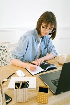 戴眼镜的女人在家庭办公桌上，一边用笔记本电脑，一边喝着咖啡写字