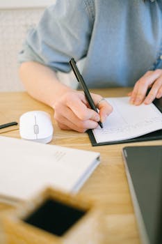 Close-up of a woman writing notes at a desk with a mouse and notebook, perfect for themes of office work and planning.