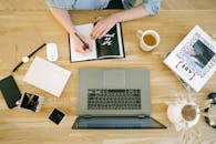 一名妇女在家庭办公桌前，拿着笔记本电脑和咖啡杯写笔记