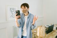一位戴着口罩的年轻女子在家庭办公环境中用智能手机进行远程通信和工作