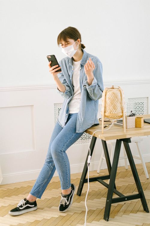Free A young woman uses her smartphone while wearing a mask in her home office. Stock Photo