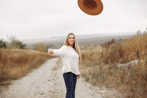 Positive young female with long blond hair in casual clothes throwing hat and smiling while standing on empty rural roadway among dry grass