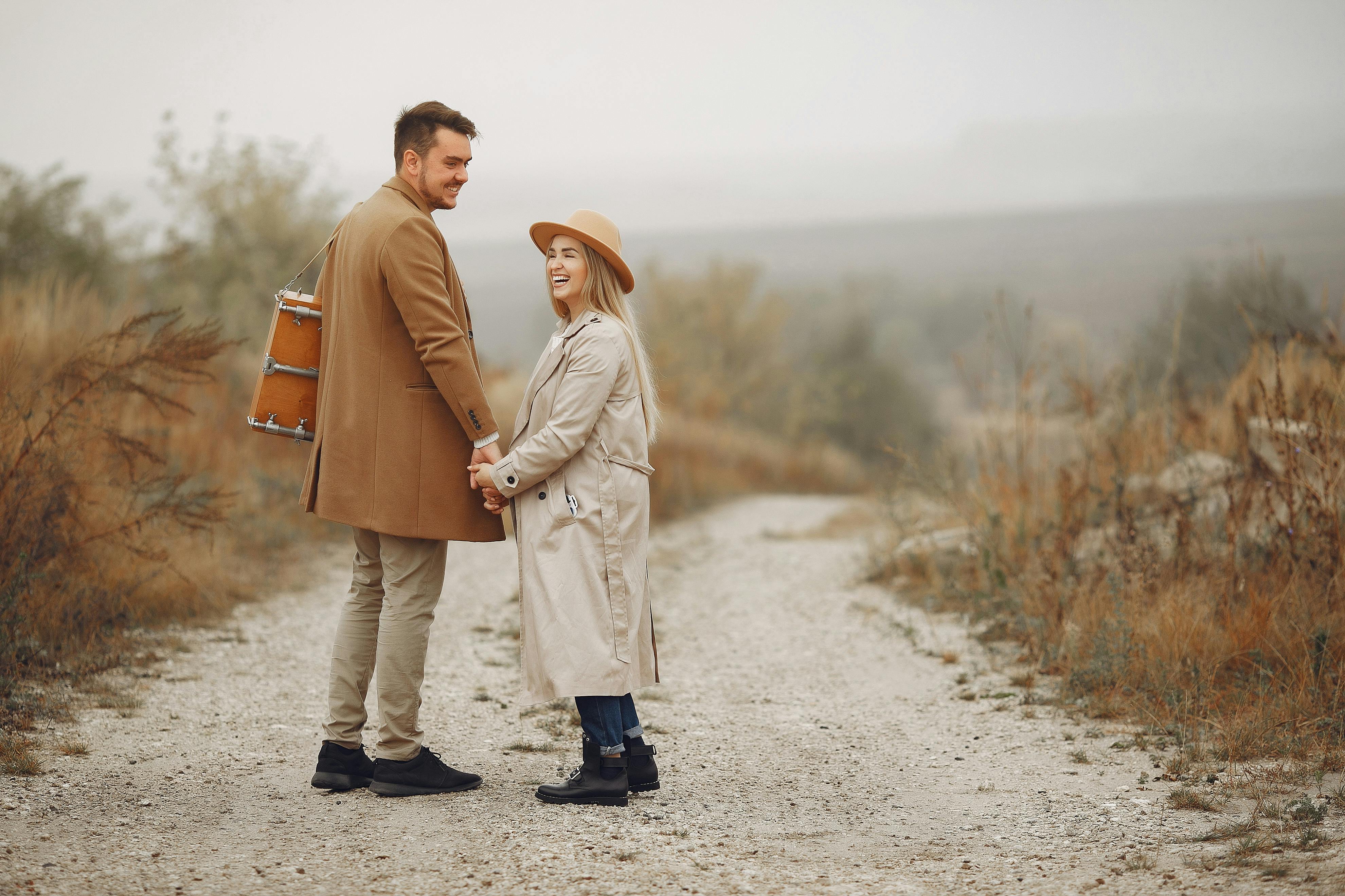 Full body of young cheerful couple in trendy clothes laughing and holding hands while walking on rural path in autumn day