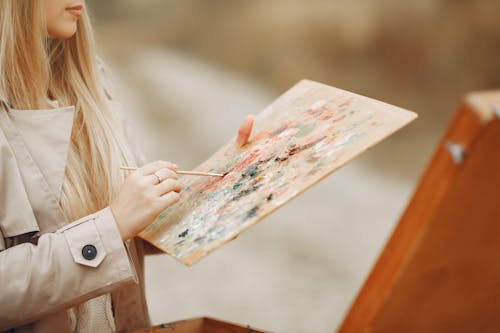 Crop unrecognizable concentrated young female artist in trench coat using paintbrush and mixing colors on palette against wooden easel