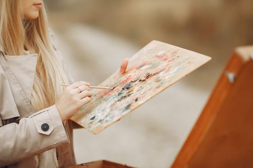 Free Crop unrecognizable concentrated young female artist in trench coat using paintbrush and mixing colors on palette against wooden easel Stock Photo