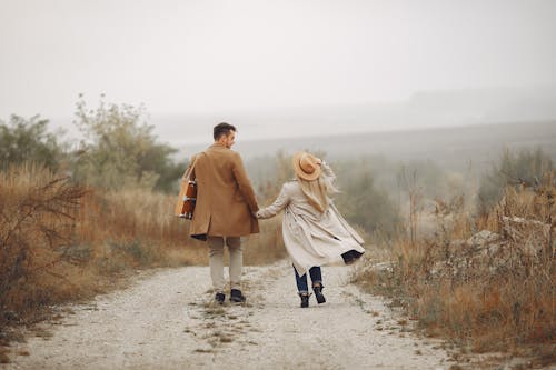 Back view of anonymous man and woman in stylish outfits holding hands and walking along rural road