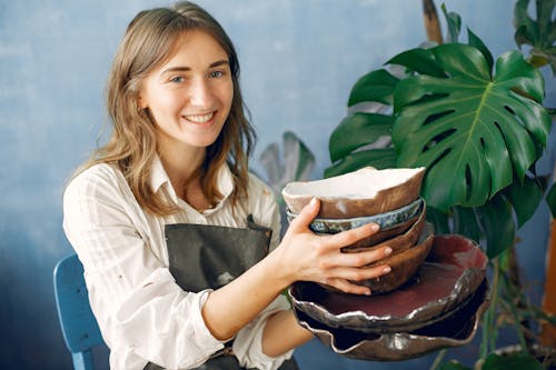 Positive smiling young craftswoman in white shirt and black apron demonstrating handmade ceramic dishware while sitting near big plant against dark blue wall in modern pottery