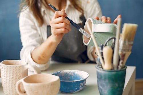 Crop anonymous craftswoman in apron painting handmade ceramic products while working in contemporary creative pottery