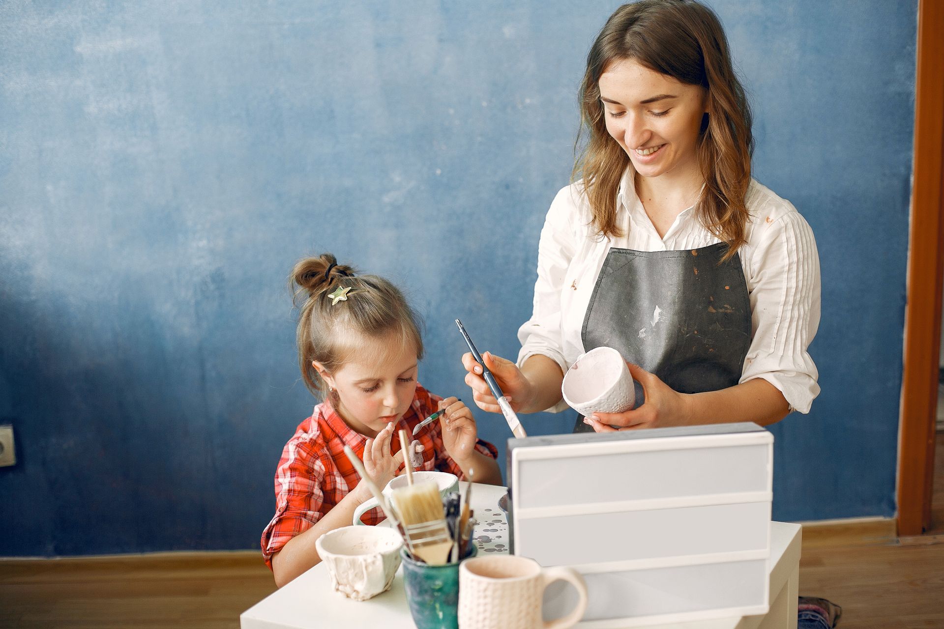Happy content female ceramist with cute little daughter painting handmade ceramic cups while working in creative workshop together against dark blue wall