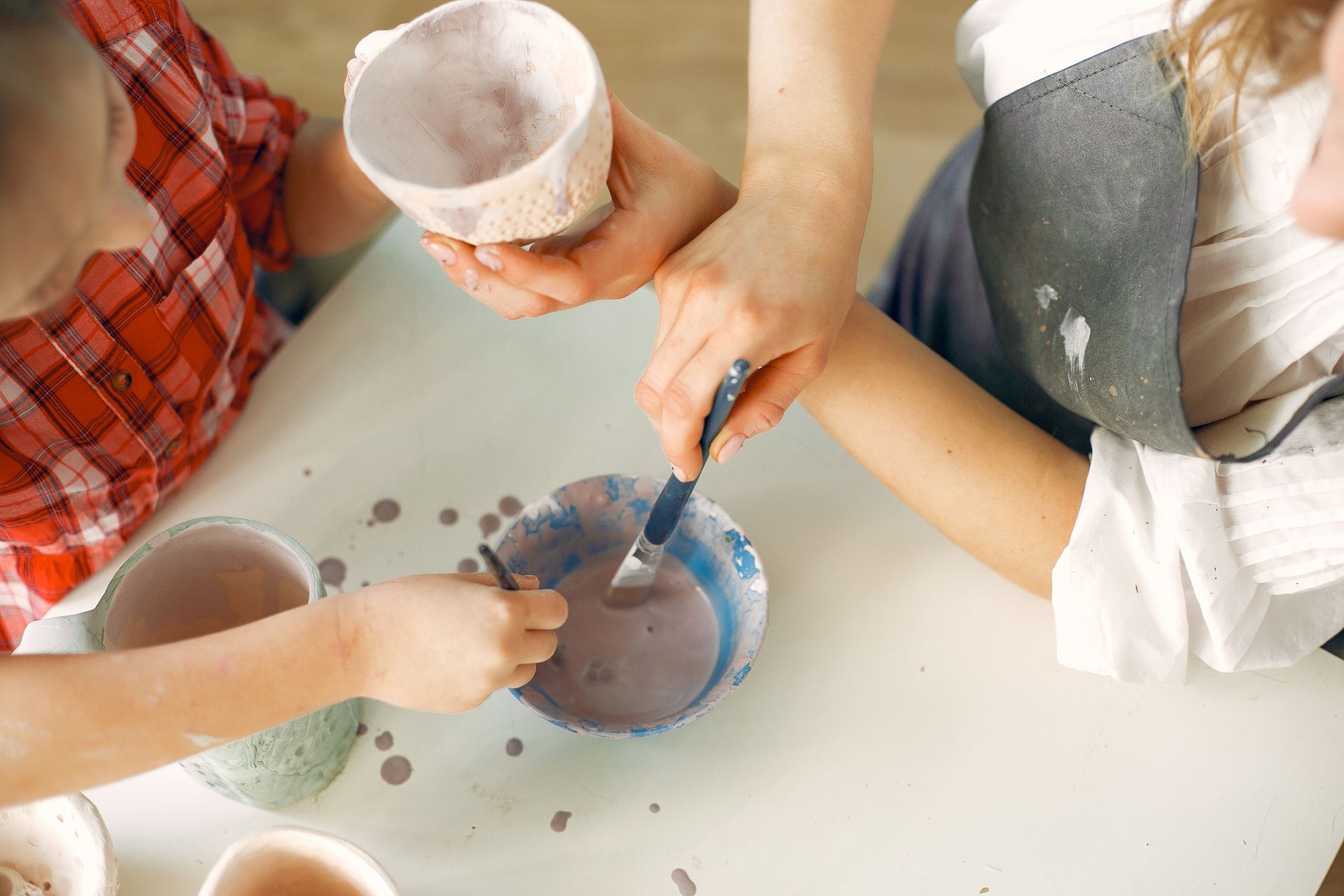 From above crop unrecognizable female ceramist and daughter in casual clothes painting handmade ceramic cups while working in creative workshop