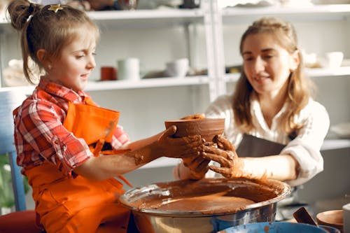 Cheerful adorable girl in casual clothes and apron making handmade ceramic bowl on pottery wheel with content mother