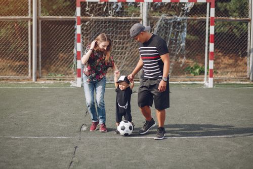 Free Full body of positive happy parents in casual clothes holding toddler son hands while walking together on football field on sunny day Stock Photo