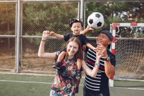 Free Positive smiling diverse parents giving piggyback ride to adorable toddler son while spending weekend together with football ball on sports ground on sunny day Stock Photo