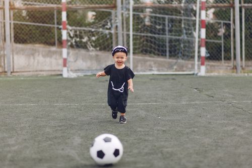 Free Full body cheerful laughing little boy in casual black outfit with ball walking on green empty sports ground on sunny summer day Stock Photo