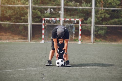 Free Full body anonymous father in black casual wear and toddler son walking with football ball on green sports ground on sunny day Stock Photo